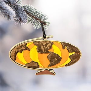 May include: A wooden globe ornament with a gold frame and a black string for hanging. The globe features brown continents and a yellow and orange background. The ornament is hanging from a snow-covered evergreen branch.