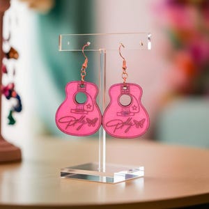 May include: A pair of bright pink guitar-shaped earrings hanging from a clear acrylic display stand. Each earring features a cutout sound hole, a small star detail, a butterfly graphic, and the cursive text Dolly engraved on the front surface.