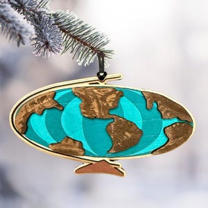 Pode incluir: Enfeite de Natal de globo terrestre de madeira com um oceano azul turquesa e continentes castanhos. O enfeite é suspenso por uma corda preta e tem uma moldura de madeira castanha clara. Pendurado em um galho de pinheiro gelado.
