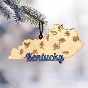 May include: Wooden Kentucky state ornament with blue lettering and various etched images. The ornament is hanging from a black string and is displayed on a frosted evergreen branch. The word "Kentucky" is in a blue, cursive font.