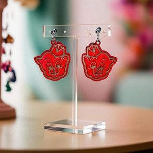 May include: A pair of red pig-shaped earrings with gold accents hanging from a clear acrylic stand.
