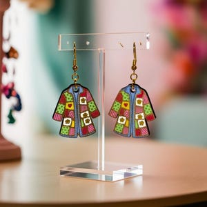 May include: A pair of dangle earrings featuring charms shaped like coats with a colorful patchwork pattern. The charms display squares in shades of green, pink, yellow, and white, accented with small flower and heart motifs, hanging from gold-toned hooks.