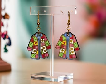 May include: A pair of dangle earrings featuring charms shaped like coats with a colorful patchwork pattern. The charms display squares in shades of green, pink, yellow, and white, accented with small flower and heart motifs, hanging from gold-toned hooks.