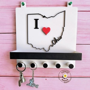 May include: White key holder with a black outline of Ohio, featuring a red heart and the word "Ohio." Includes a shelf and five key hooks. Two keys and a keychain are hanging from the hooks. The key holder is mounted on a pink wooden surface.