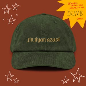 Op de afbeelding: Een donkergroene corduroy baseballcap met de geborduurde tekst "jin jiyan azadi" in goud. De cap heeft een gebogen klep en een ontwerp met zes panelen. De achtergrond is roodbruin met witte ster-doodles en de tekst "ORIGINAL DESIGN, ONLY AVAILABLE AT THE DUMB SHOP!!!".