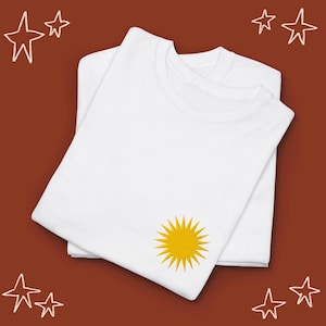 Op de afbeelding: Twee witte T-shirts die op elkaar gevouwen zijn, een met een geborduurd geel zonontwerp op de voorkant.