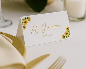 May include: A white place card with gold script reading "Lily Jameson" and sunflower illustrations. The card sits on a white tablecloth, with gold cutlery and a folded napkin. A lit candle and floral arrangement are in the background.