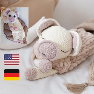 Könnte beinhalten: Ein gehäkeltes Schweinchen-Plüschtier mit weißem Körper und rosa Ohren und Schnauze. Das Schweinchen liegt auf einer weißen Decke mit einer braunen Decke und einem braunen und weißen Kissen dahinter.