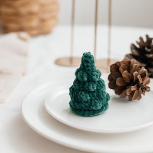 May include: A small, dark green, crocheted Christmas tree sits on a white plate. Two pine cones and gold candle holders are in the background. The tree is made of a soft, textured yarn and is a festive decoration.