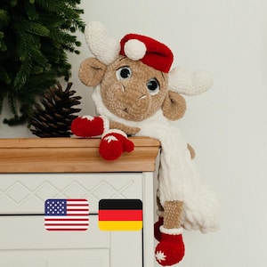 Puede incluir: Un juguete de reno de crochet marrón que lleva un gorro de Papá Noel rojo y una bufanda blanca. El reno está sentado en un estante de madera con un árbol de Navidad en el fondo.