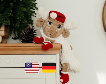 May include: A brown crocheted reindeer toy wearing a red Santa hat and a white scarf. The reindeer is sitting on a wooden shelf with a Christmas tree in the background.