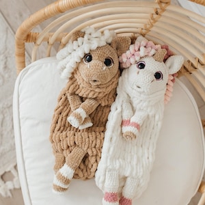 May include: Two handmade plush animal toys. One is tan with white and tan striped accents, the other is white with pink and white striped accents. Both have large black eyes and curly hair. They are lying on a white cushion.