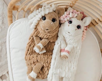 May include: Two handmade plush animal toys. One is tan with white and tan striped accents, the other is white with pink and white striped accents. Both have large black eyes and curly hair. They are lying on a white cushion.