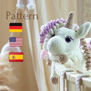 May include: A white crocheted unicorn toy with pink and brown yarn accents. The unicorn is sitting on a white crib rail. The text "Pattern" is displayed above the unicorn, with flags of Germany, the United States, and Spain below the text.