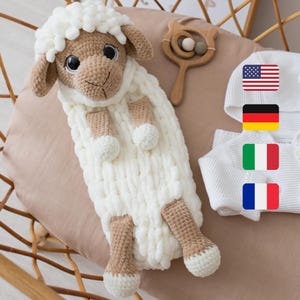 May include: A white crocheted lamb stuffed animal with brown eyes and a white knitted blanket. The lamb is lying in a wicker basket. There are flags of the United States, Germany, Italy, and France in the background.