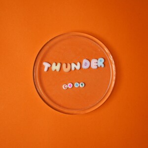 May include: A clear, circular coaster with the words "THUNDER CONT" spelled out in colorful, beaded letters. The coaster sits on a vibrant orange background, creating a playful and eye-catching design.