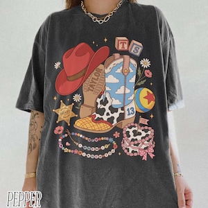 May include: A charcoal grey t-shirt featuring a graphic print of a red cowboy hat, a blue and white cloud-patterned cowboy boot with a cow print section, a yellow and red ball, a sheriff star, alphabet blocks, and a beaded necklace with text.