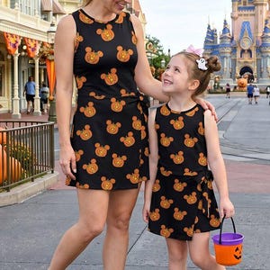 May include: Two sleeveless black dresses featuring an all-over orange pumpkin pattern shaped like mouse ears. Each dress has a wrap-style skirt with a side tie detail. The dresses are paired with light-colored sneakers and a small orange pumpkin bucket.