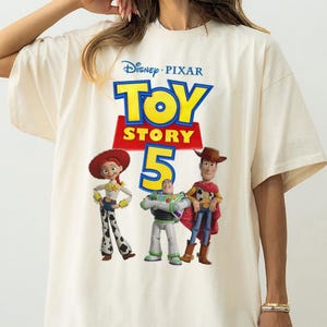 Op de afbeelding: Een off-white t-shirt met korte mouwen en een grafische print van het Disney Pixar Toy Story 5-logo, met de personages Jessie, Buzz Lightyear en Woody onder de tekst. Het ontwerp is gecentreerd op de voorzijde van het kledingstuk.