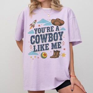 May include: A light purple short-sleeve t-shirt featuring a graphic print with the text "YOU'RE A COWBOY LIKE ME" in dark blue letters. The design includes a brown cowboy hat, a rocket, a yellow and blue ball with a red star, and brown boots.