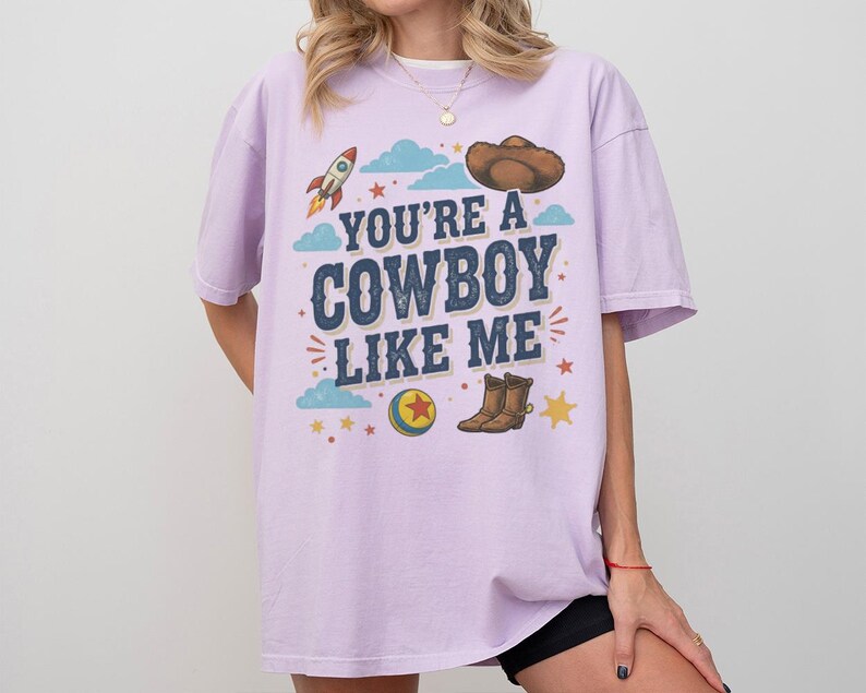May include: A light purple short-sleeve t-shirt featuring a graphic print with the text "YOU'RE A COWBOY LIKE ME" in dark blue letters. The design includes a brown cowboy hat, a rocket, a yellow and blue ball with a red star, and brown boots.