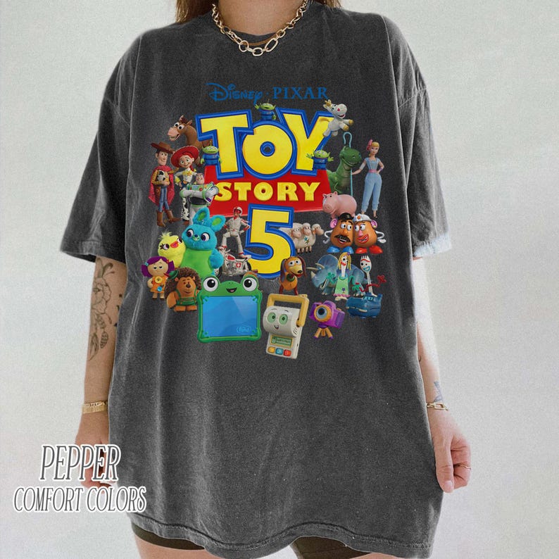 May include: A charcoal gray short-sleeve t-shirt featuring a large graphic print of various Toy Story characters surrounding the yellow and red text Toy Story 5. The shirt has a crew neckline and a relaxed fit with a vintage-style washed fabric finish.