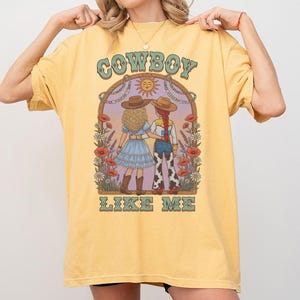 Puede incluir: Una camiseta de color amarillo p&aacute;lido con un estampado gr&aacute;fico de dos figuras con sombreros de vaquero de pie una al lado de la otra. El dise&ntilde;o incluye elementos florales, un icono de sol y el texto "COWBOY LIKE ME" en una fuente estilizada.