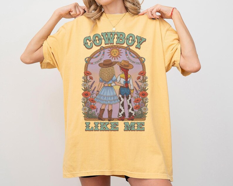 Puede incluir: Una camiseta de color amarillo p&aacute;lido con un estampado gr&aacute;fico de dos figuras con sombreros de vaquero de pie una al lado de la otra. El dise&ntilde;o incluye elementos florales, un icono de sol y el texto "COWBOY LIKE ME" en una fuente estilizada.