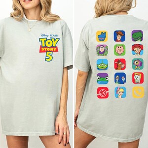 May include: A light gray short-sleeve t-shirt featuring the Disney Pixar Toy Story 5 logo on the front chest. The back of the shirt displays a grid of fifteen colorful square icons, each containing a different character or symbol from the film series.