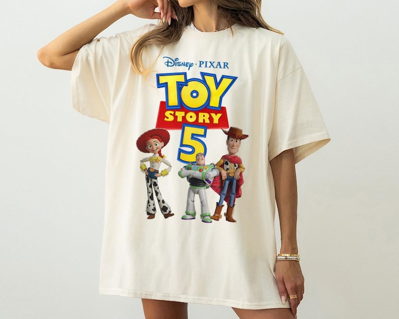 Op de afbeelding: Een off-white t-shirt met korte mouwen en een grafische print van het Disney Pixar Toy Story 5-logo, met de personages Jessie, Buzz Lightyear en Woody onder de tekst. Het ontwerp is gecentreerd op de voorzijde van het kledingstuk.