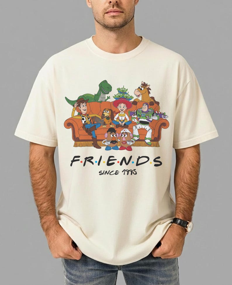 K&ouml;nnte beinhalten: Ein cremefarbenes Kurzarm-T-Shirt mit einer Grafik von Toy Story-Figuren auf einem orangefarbenen Sofa. Unter den Figuren steht der Text F.R.I.E.N.D.S und SINCE 1995 in schwarzer Schrift mit bunten Punkten zwischen den Buchstaben.