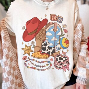 May include: A cream-coloured t-shirt featuring a graphic print of a red cowboy hat, a cow-print cowboy boot with a blue sky and cloud pattern, a sheriff star, a colourful ball, a beaded necklace with the text I KNEW IT, and a cake with a flag.