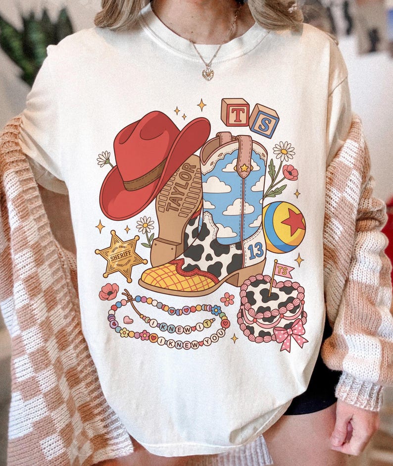 May include: A cream-coloured t-shirt featuring a graphic print of a red cowboy hat, a cow-print cowboy boot with a blue sky and cloud pattern, a sheriff star, a colourful ball, a beaded necklace with the text I KNEW IT, and a cake with a flag.