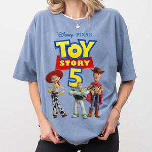 Op de afbeelding: Een lichtblauw t-shirt met een grafische print van het Disney Pixar Toy Story 5 logo in gele en rode tekst, vergezeld van illustraties van de personages Jessie, Buzz Lightyear en Woody die in een rij onder de titel staan.