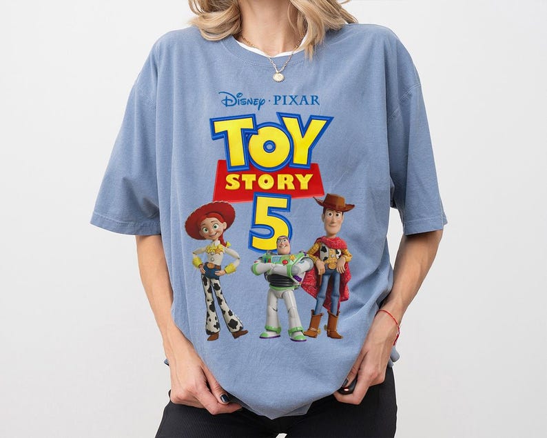 Op de afbeelding: Een lichtblauw t-shirt met een grafische print van het Disney Pixar Toy Story 5 logo in gele en rode tekst, vergezeld van illustraties van de personages Jessie, Buzz Lightyear en Woody die in een rij onder de titel staan.