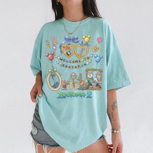 May include: Light blue t-shirt featuring a colorful graphic from the movie Zootopia. The design includes cartoon characters, hearts, balloons, and the text "WELCOME TO ZOOTOPIA" and "Zootopia 2".