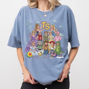May include: A dusty blue short-sleeve t-shirt featuring a graphic print of multiple characters dressed in various outfits, including a cowboy, a space ranger, and a dinosaur. The text TS is printed in yellow at the top center of the graphic design.