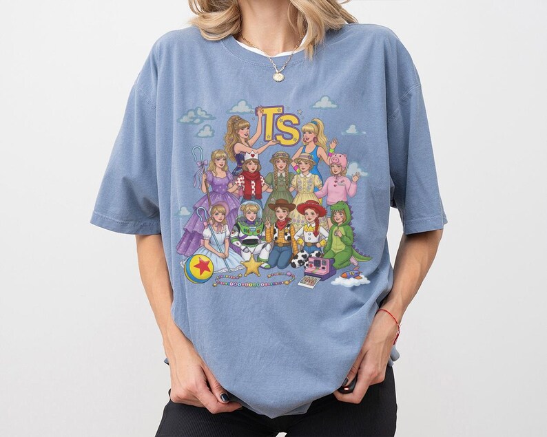 May include: A dusty blue short-sleeve t-shirt featuring a graphic print of multiple characters dressed in various outfits, including a cowboy, a space ranger, and a dinosaur. The text TS is printed in yellow at the top center of the graphic design.