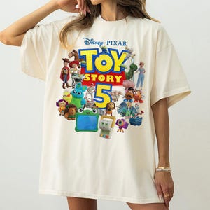 May include: An off-white short-sleeve t-shirt featuring a large, colorful graphic print on the front. The design displays the Disney Pixar Toy Story 5 logo in bold yellow and red text, surrounded by a collection of various animated character figures.