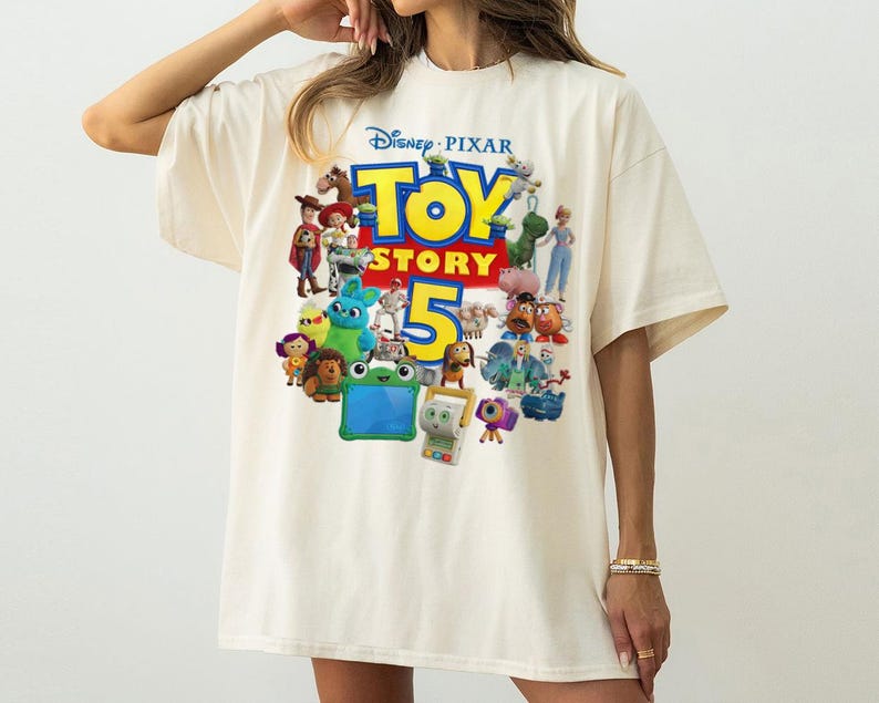 May include: An off-white short-sleeve t-shirt featuring a large, colorful graphic print on the front. The design displays the Disney Pixar Toy Story 5 logo in bold yellow and red text, surrounded by a collection of various animated character figures.