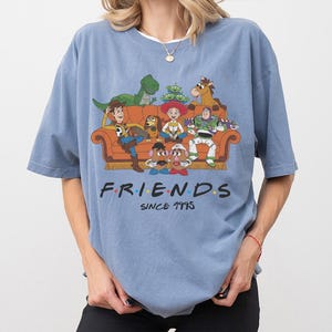K&ouml;nnte beinhalten: Ein staubblaues Kurzarm-T-Shirt mit einer Grafik von Toy Story-Figuren, die auf einem Sofa sitzen. Der Text FRIENDS SINCE 1995 ist in schwarzer Schrift gedruckt. Zu sehen sind Woody, Buzz Lightyear, Jessie, Rex, Slinky Dog, Mr. und Mrs. Potato Head sowie die Aliens.