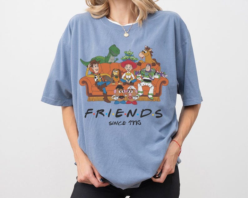 K&ouml;nnte beinhalten: Ein staubblaues Kurzarm-T-Shirt mit einer Grafik von Toy Story-Figuren, die auf einem Sofa sitzen. Der Text FRIENDS SINCE 1995 ist in schwarzer Schrift gedruckt. Zu sehen sind Woody, Buzz Lightyear, Jessie, Rex, Slinky Dog, Mr. und Mrs. Potato Head sowie die Aliens.