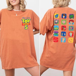 May include: A burnt orange short-sleeve t-shirt featuring the Disney Pixar Toy Story 5 logo on the front left chest. The back of the shirt displays a grid of fifteen colorful square icons, each depicting a different character from the Toy Story franchise.