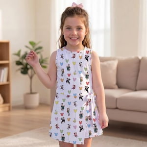 May include: A sleeveless white dress featuring an all-over print of cartoon characters holding colorful balloons shaped like mouse ears. The dress has a wrap-style skirt with a side tie detail at the waist, creating a layered appearance.