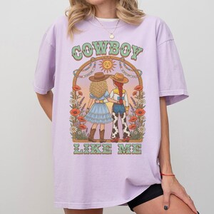 Puede incluir: Una camiseta de color morado claro con un estampado gr&aacute;fico de dos figuras con sombreros de vaquero y ropa del oeste bajo un arco con flores y un sol. El texto "COWBOY" aparece sobre las figuras y "LIKE ME" est&aacute; impreso debajo.