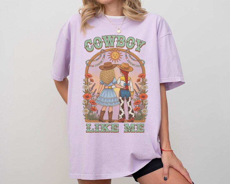 Puede incluir: Una camiseta de color morado claro con un estampado gr&aacute;fico de dos figuras con sombreros de vaquero y ropa del oeste bajo un arco con flores y un sol. El texto "COWBOY" aparece sobre las figuras y "LIKE ME" est&aacute; impreso debajo.