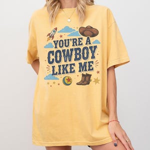 May include: A yellow short-sleeve t-shirt featuring a graphic print with the text "YOU'RE A COWBOY LIKE ME" in blue letters. The design includes a rocket, a brown cowboy hat, a colourful ball with a red star, cowboy boots, clouds, and stars.