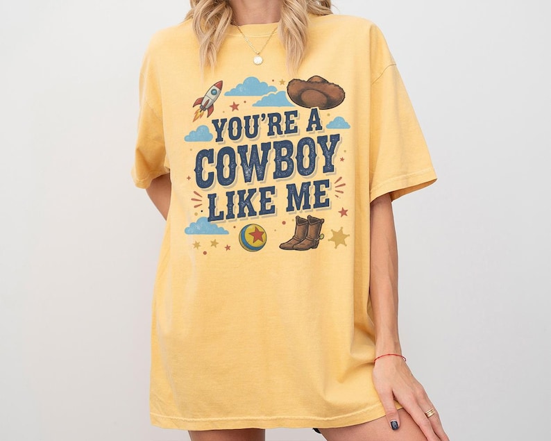 May include: A yellow short-sleeve t-shirt featuring a graphic print with the text "YOU'RE A COWBOY LIKE ME" in blue letters. The design includes a rocket, a brown cowboy hat, a colourful ball with a red star, cowboy boots, clouds, and stars.