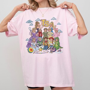 May include: A light pink t-shirt featuring a colorful graphic print of various animated characters dressed in costumes. The design includes the yellow letters TS at the top, a blue and yellow ball, a star, and a pink toy camera on a light blue background.