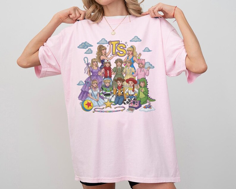 May include: A light pink t-shirt featuring a colorful graphic print of various animated characters dressed in costumes. The design includes the yellow letters TS at the top, a blue and yellow ball, a star, and a pink toy camera on a light blue background.